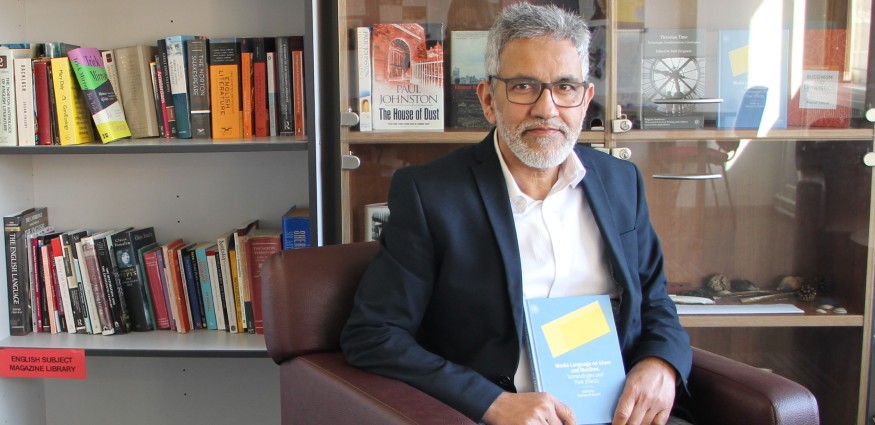 A male wearing a white shirt and navy blue blazer sits in a chair holding a book.